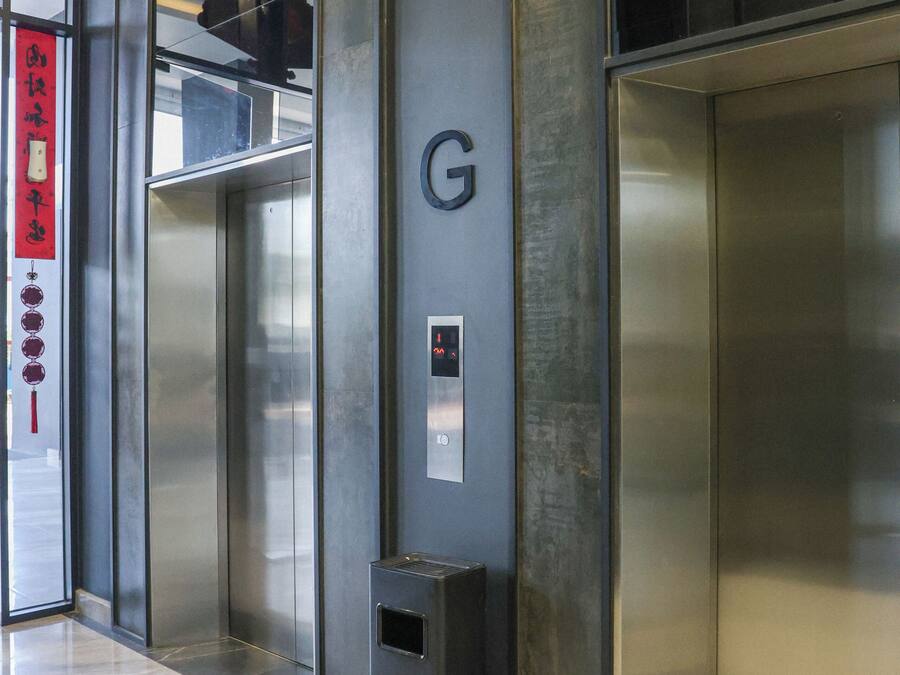 Een lift met een metalen afwerking, een aan de muur bevestigd noodtelefoon en een prullenbak.