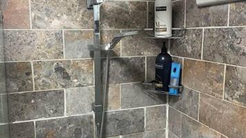A shower with a handheld showerhead, a bottle of shampoo, and a soap dispenser on a shelf.