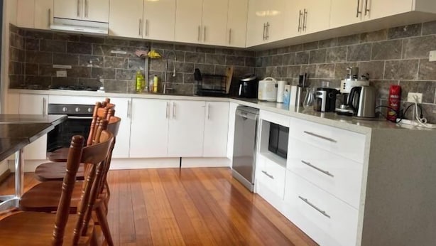 A modern kitchen with white cabinets, a wooden dining table, and a stone backsplash. - 3 Bed room Villa 19 Mins to Melbourne center (Laverton)