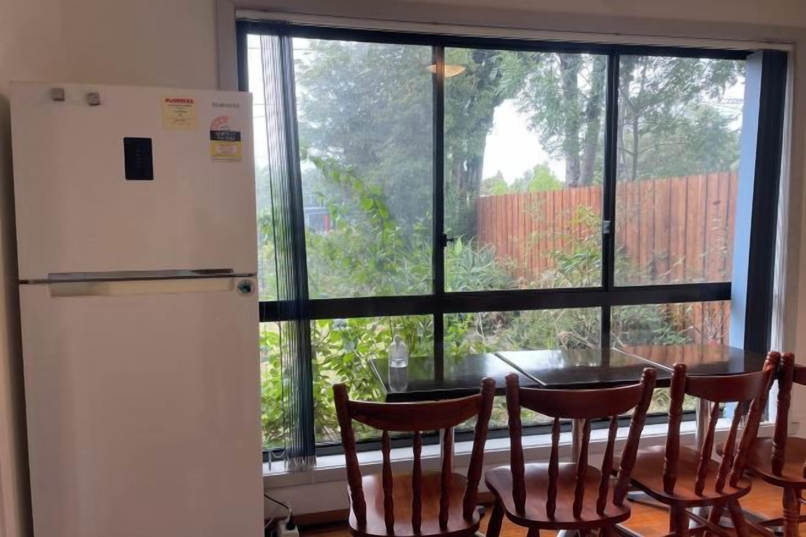 A kitchen with a refrigerator, a dining table with chairs, and a large window with a view of a wooden fence and greenery.