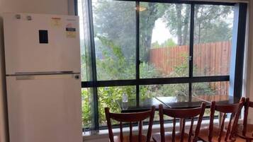 A kitchen with a refrigerator, a dining table with chairs, and a large window with a view of a wooden fence and greenery.