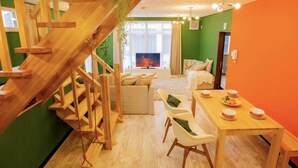 A modern living room with a wooden staircase, a sofa set, a dining table with plates, and a TV in the background. - Yuzawa Onsen Lodge House B (Minamiuonuma)