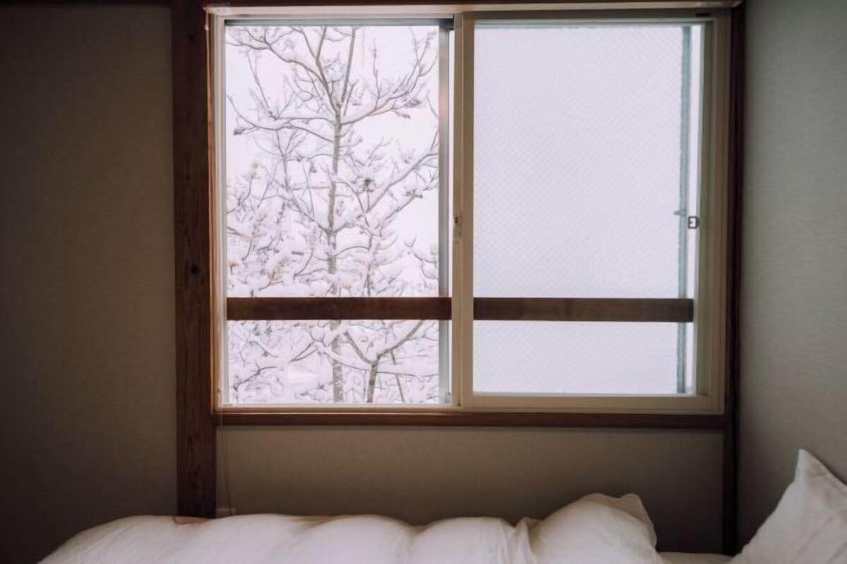 Una habitación con una cama, un ventanal con vistas a árboles desnudos y una puerta.