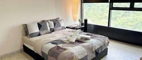 A modern bedroom with a large bed, geometric patterned bedding, a bedside lamp, and a view of greenery through a large window.