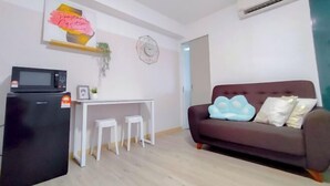 Un espacio de estar con un refrigerador negro, un escritorio blanco, dos taburetes blancos, un sofá marrón oscuro con almohadones, un reloj de pared y un equipo de aire acondicionado.
