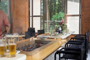 A restaurant with a grill, tables, chairs, and a view of greenery outside. - Nasu Prosperity Guesthouse (Nasu)
