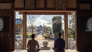 Two individuals meditating in a room with a view of a traditional Japanese garden and a building with a blue roof. - TEMPLESTAY ZENSŌ (Chiyoda)