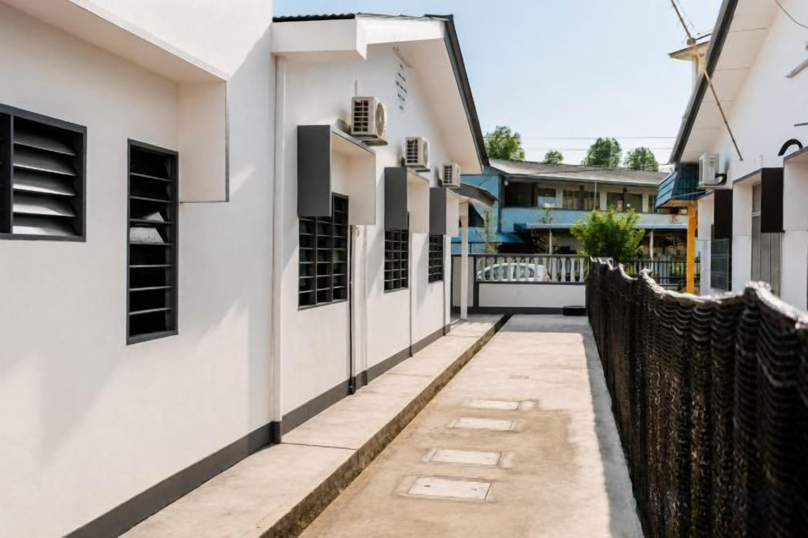 Un étroit sentier mène à un bâtiment doté d’une entrée fermée par un portail, de climatiseurs et qui se trouve sous un ciel dégagé.