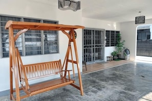 Et værelse med gyngestol i træ, store vinduer, en potteplante og udsigt til en bygning.