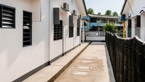 A narrow pathway between buildings with a concrete floor, a black fence on one side, and a blue building in the background.