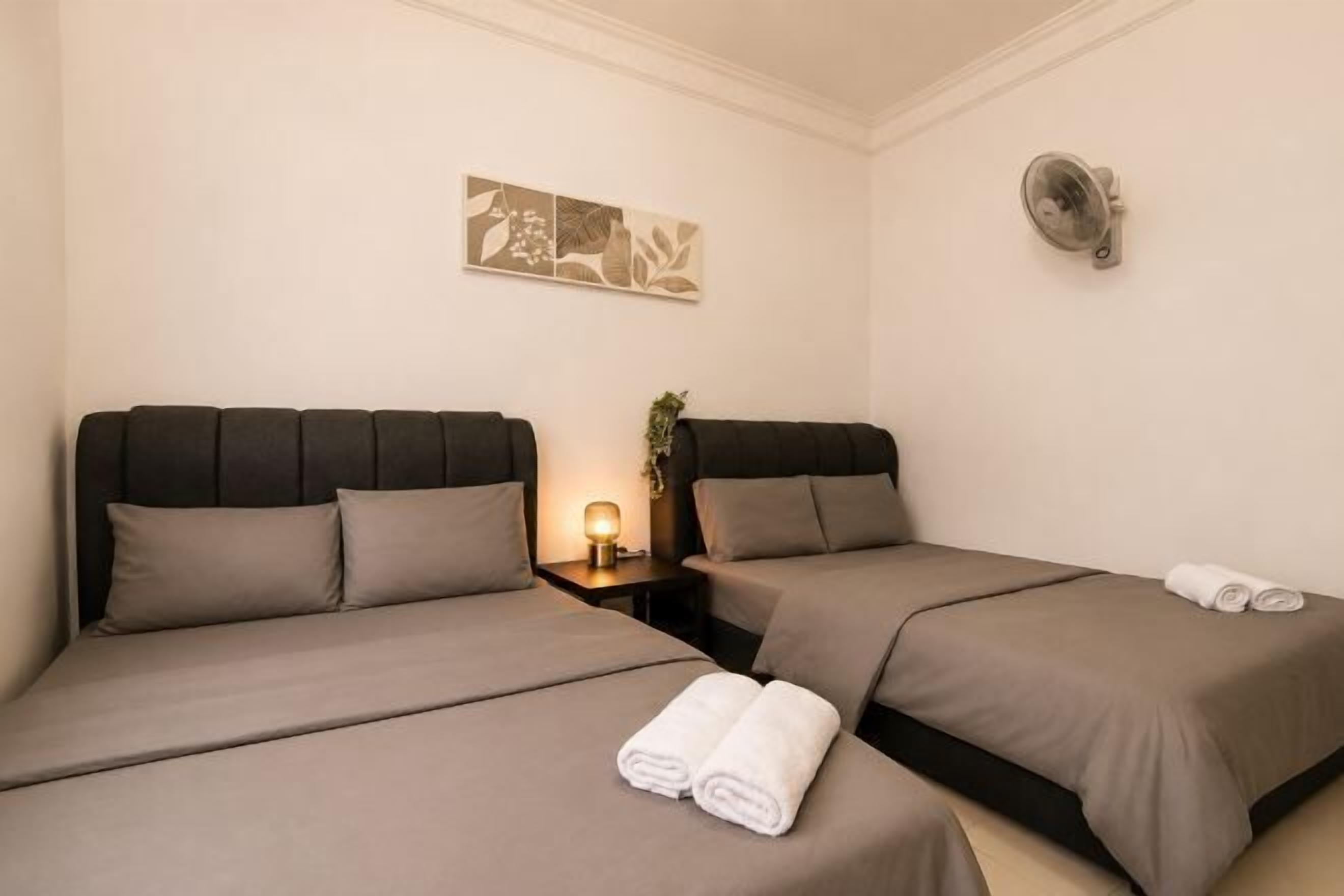 A hotel room with two beds, a bedside table with a lamp, and a wall-mounted fan.