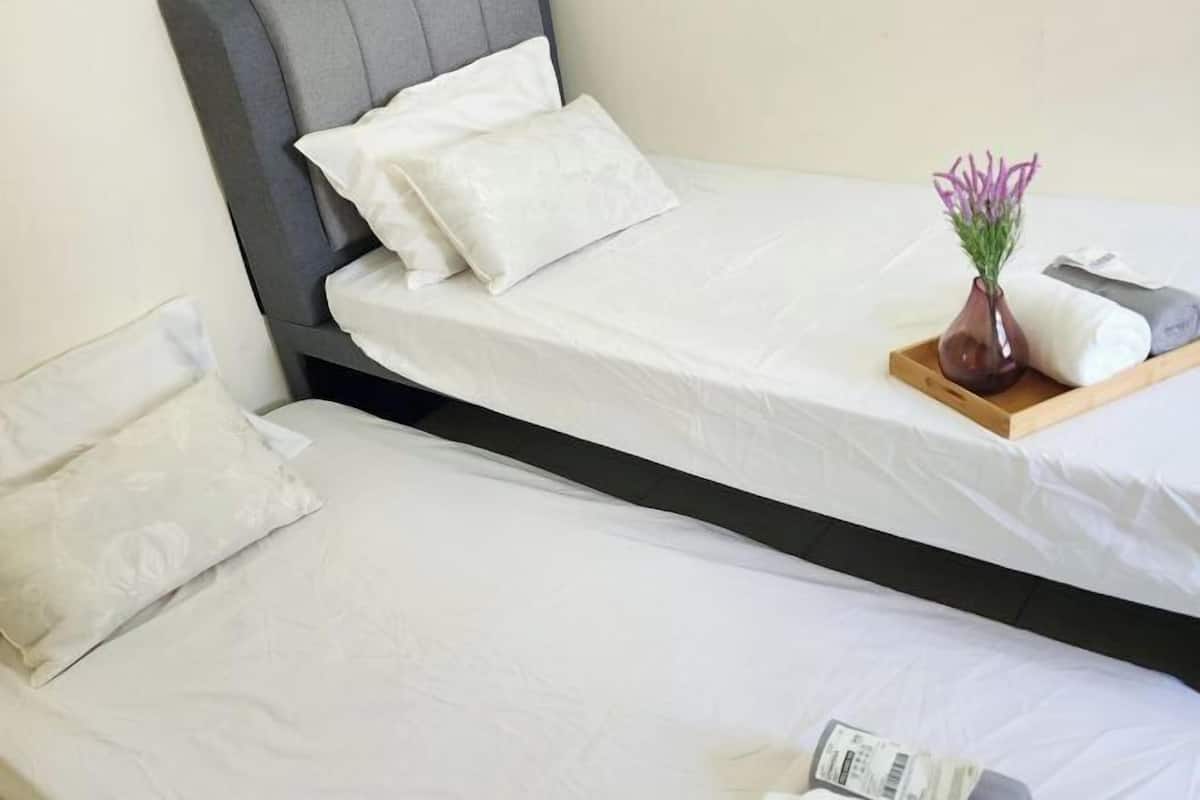 Uma cama de casal com roupa de cama branca, cabeceira cinza, um vaso com flores e uma bandeja com um travesseiro e uma toalha.