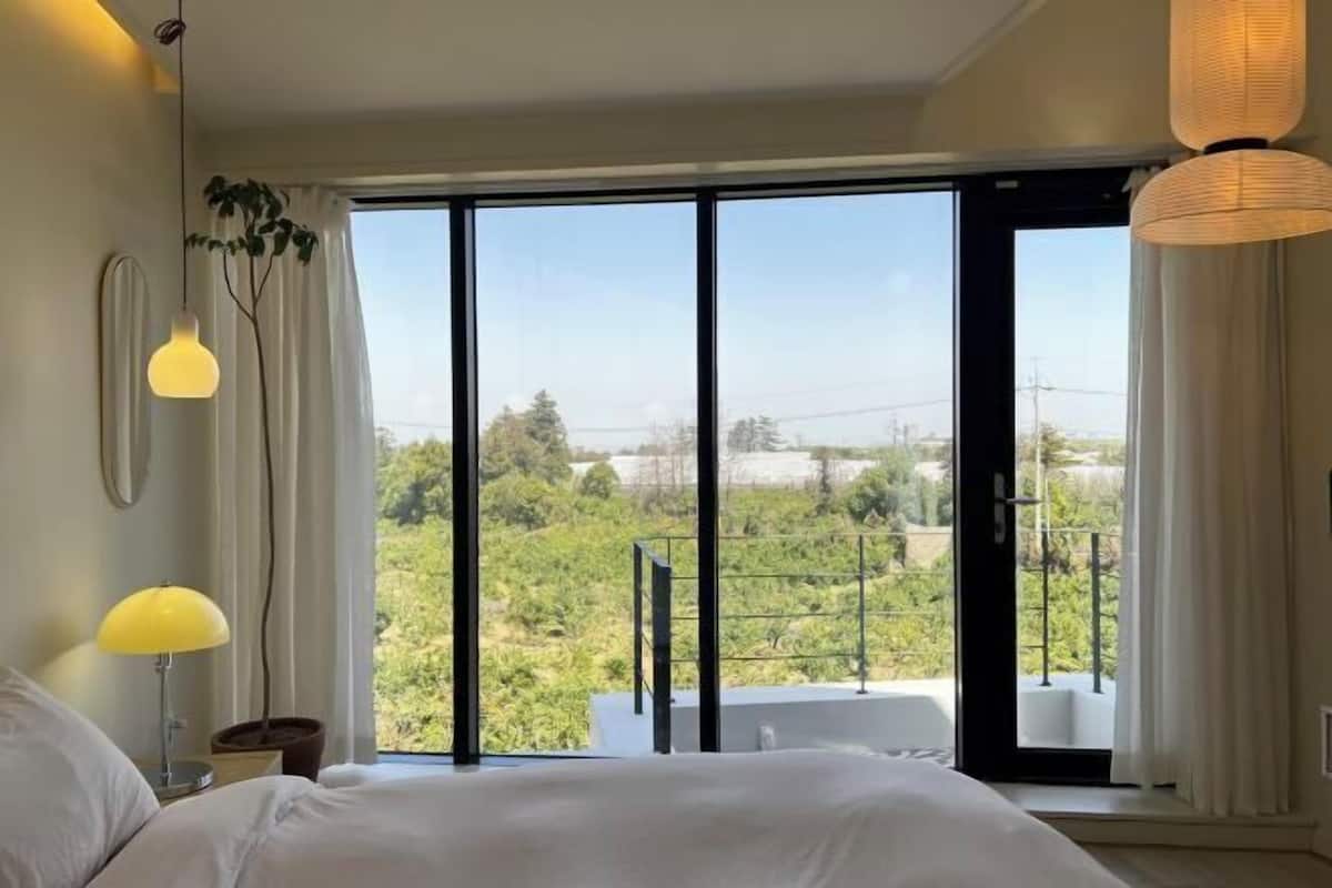 A bedroom with a large bed, a nightstand, a lamp, and a view of greenery through a sliding glass door.
