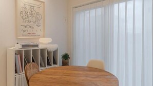 A modern dining area with a round wooden table, white chairs, a white shelf with books, a framed artwork, and a large window with sheer curtains. - Mon Monde Aewol (Jeju)