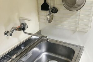 A stainless steel kitchen sink with a faucet, a soap dispenser, and a strainer rack with kitchen utensils. - Guesthouse Atelier Ten (Osaka)