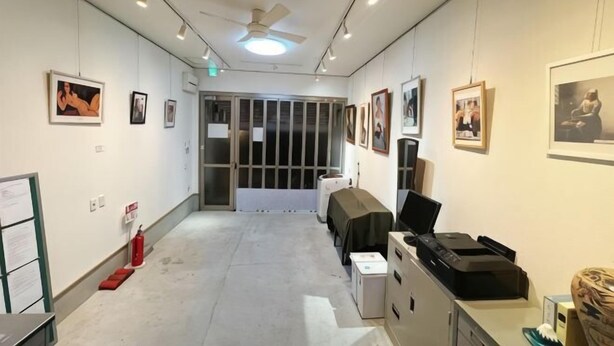 A hallway with a ceiling fan, framed pictures on the walls, a fire extinguisher, and a table with a laptop. - Guesthouse Atelier Ten (Osaka)