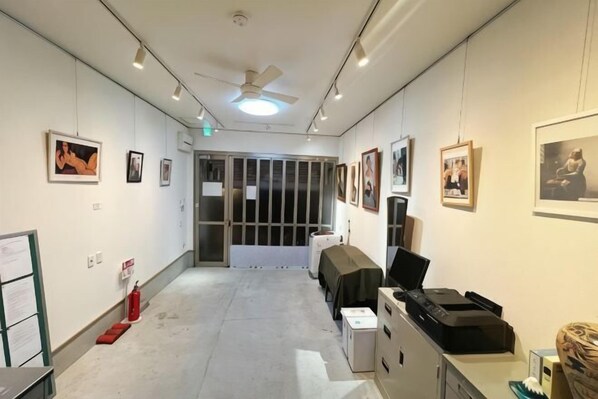 A hallway with a ceiling fan, framed pictures on the walls, a fire extinguisher, and a table with a laptop. - Guesthouse Atelier Ten (Osaka)
