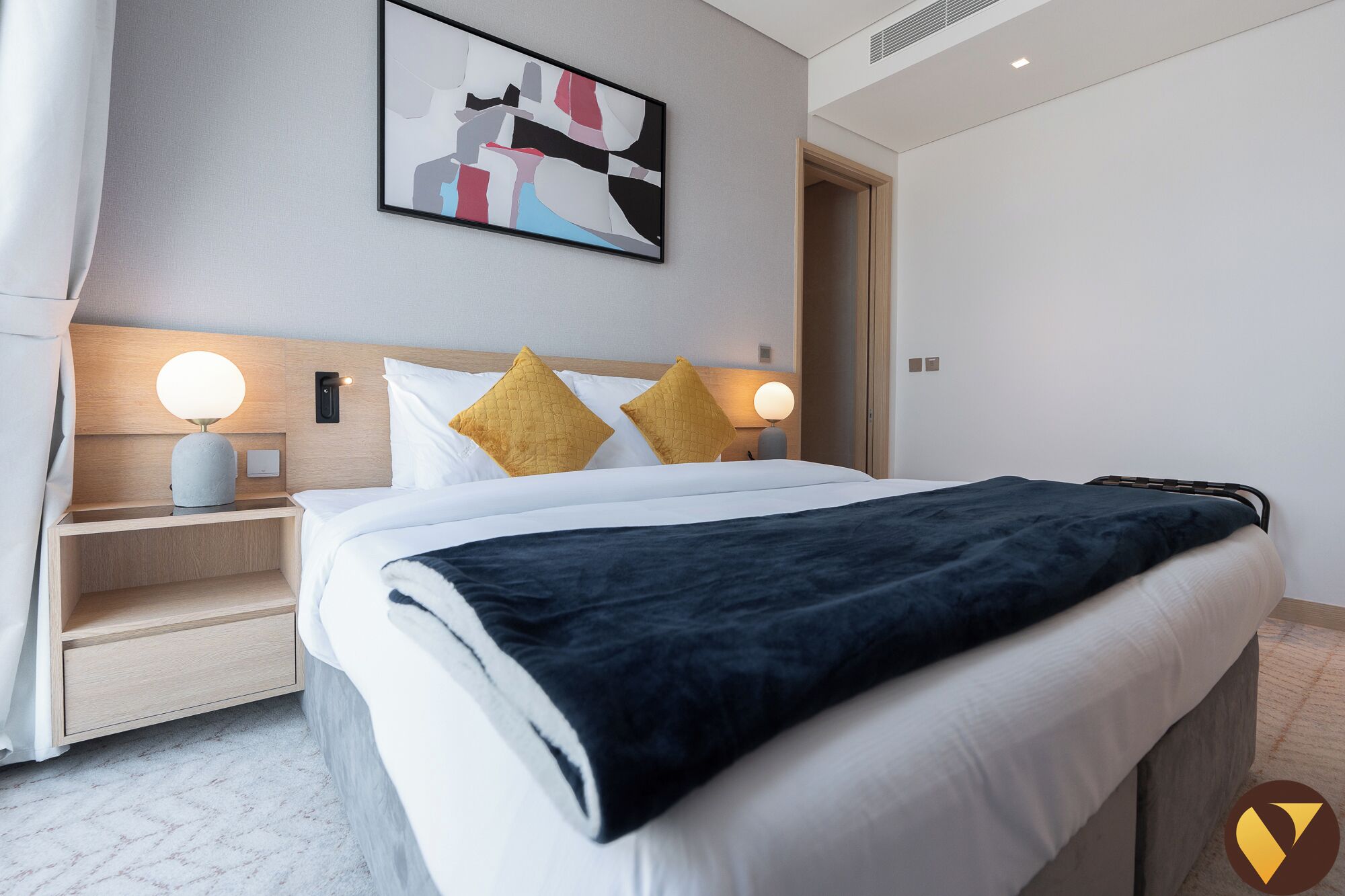 A modern hotel room with a neatly made bed, two bedside tables with lamps, a headboard, and a wall-mounted abstract painting.
