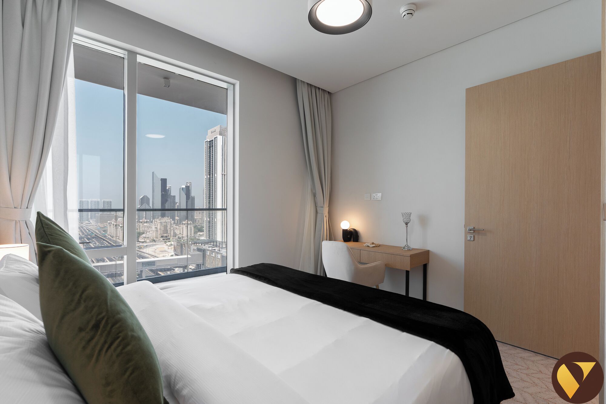 Habitación de hotel con una cama grande, un escritorio con lámpara, vista a la ciudad y una puerta.