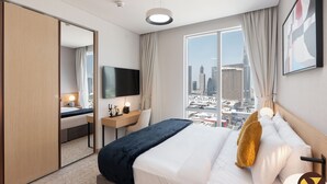 Habitación de hotel con cama, escritorio con silla, televisor y ventana grande con vista a la ciudad.