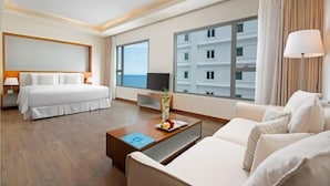 A hotel room with a bed, a sofa, a coffee table, a TV, and large windows offering a view of a building. - A La Carte Beachfront Lovers Bliss Retreat Studio (Da Nang)