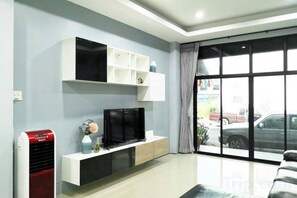 A living room with a flat-screen TV, a white entertainment unit, a red heater, and a large window with a view of the street. - Khoksametchun Hostel Plus 1 (Hat Yai)