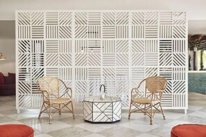 A room with a geometric patterned partition, two wicker chairs, a round table with a geometric design, and a red ottoman. - Paradise Cove Boutique Hotel (Mauritius)