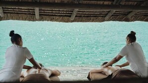 Deux personnes sont assises sur un banc en bois, sous un toit de chaume, et font face à une étendue d’eau.