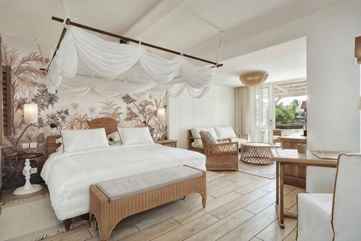 Een slaapkamer met een groot bed, een rieten bankje, een bureau en uitzicht op het groen.