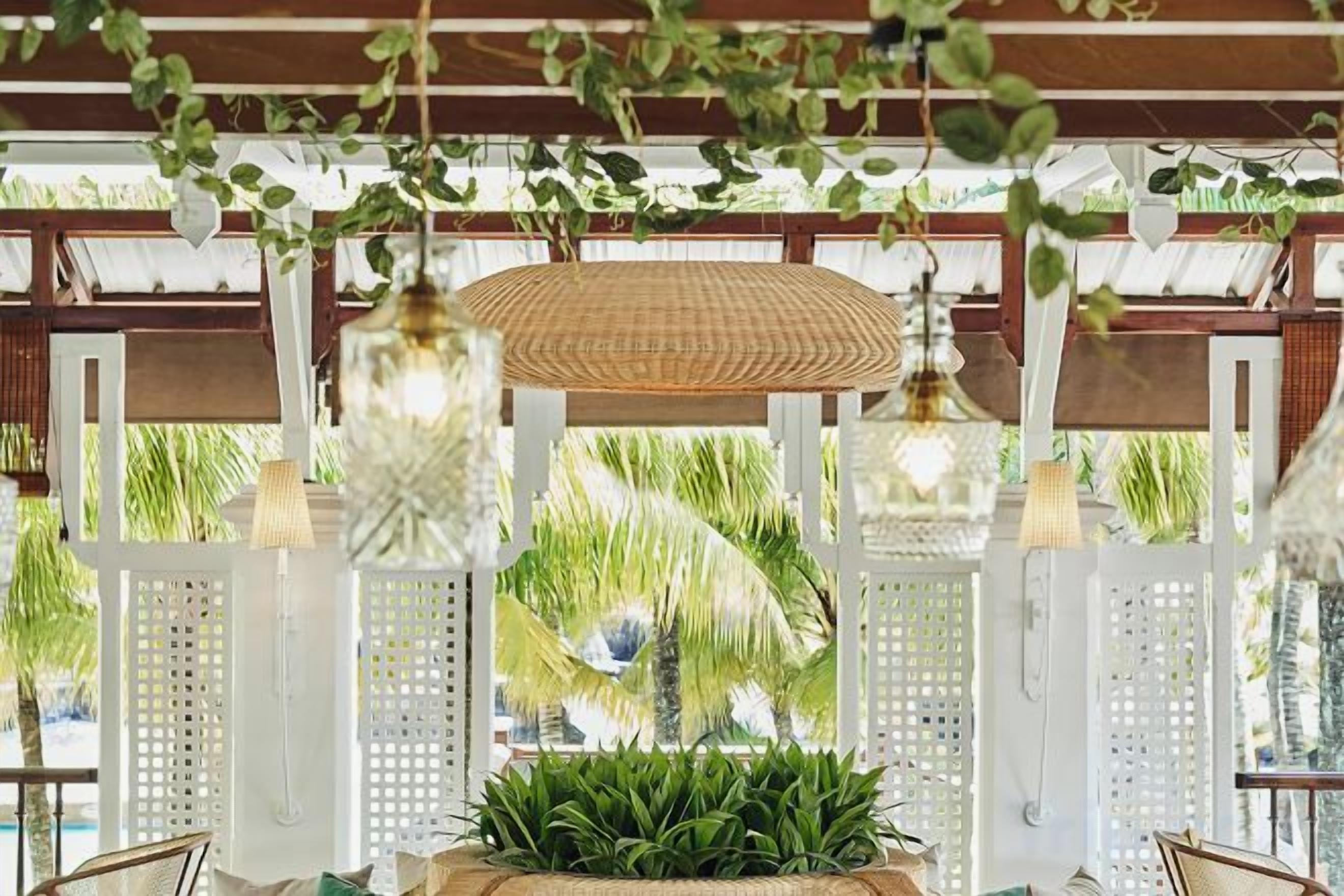 A covered outdoor seating area with hanging lights, a woven ceiling, and a view of palm trees.