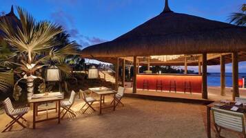 Zona de bar en la playa con techo de paja, asientos al aire libre y vista al océano.