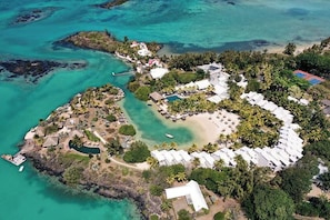 Vue aérienne d’un complexe hôtelier tropical situé sur une île, avec une plage, des palmiers et un ensemble de bâtiments blancs.