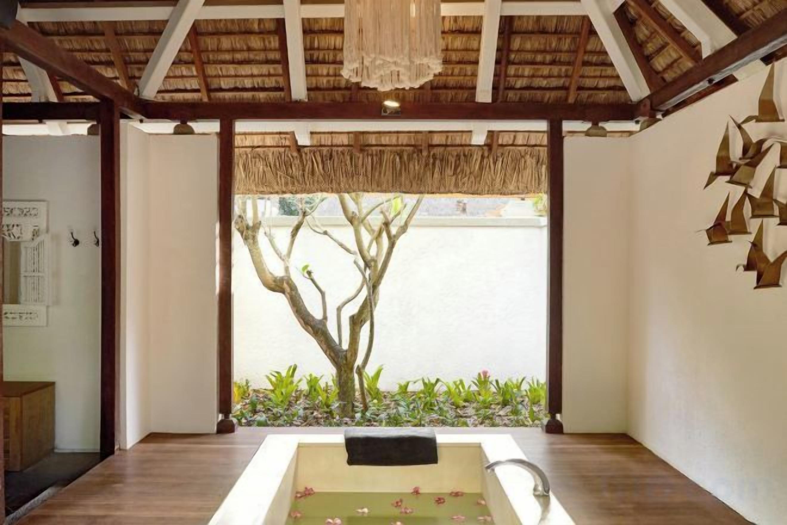 A room with a wooden floor, a bathtub with a towel, a tree outside, and a thatched roof.