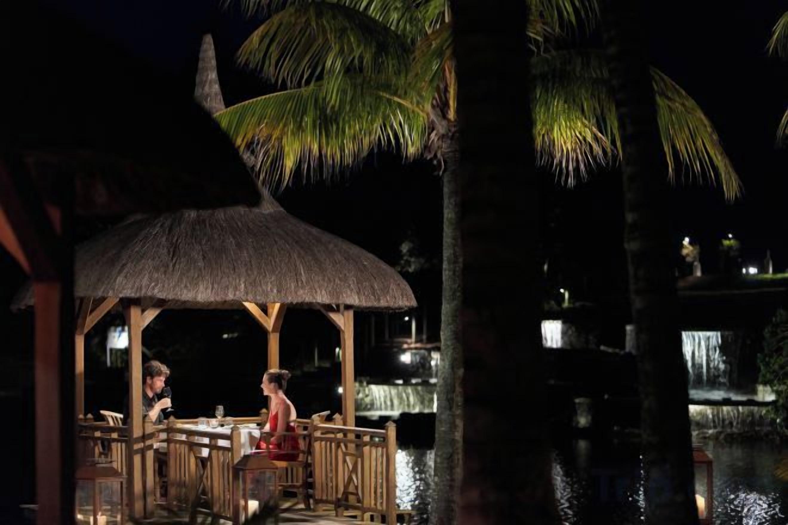 A thatched-roof outdoor dining area with wooden furniture, overlooking a body of water.