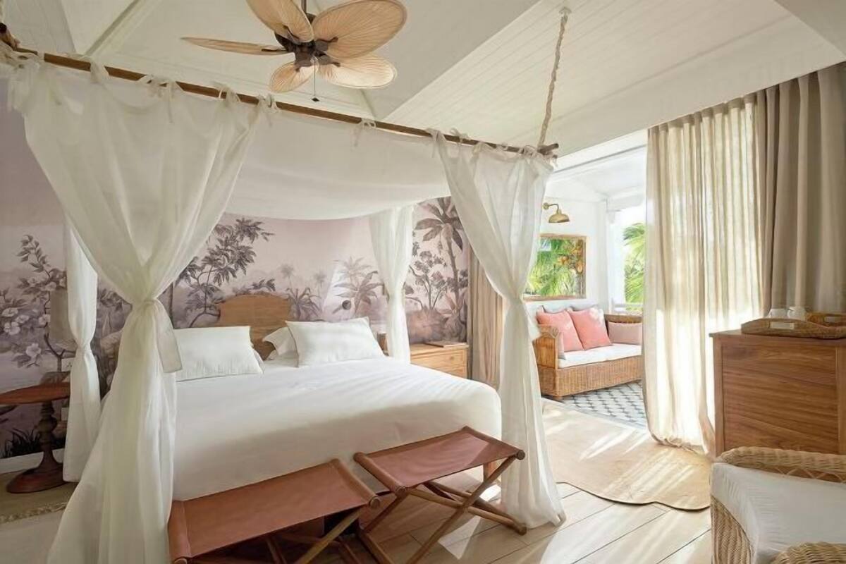 Een slaapkamer met hemelbed, plafondventilator, een houten bankje en uitzicht op de omgeving.
