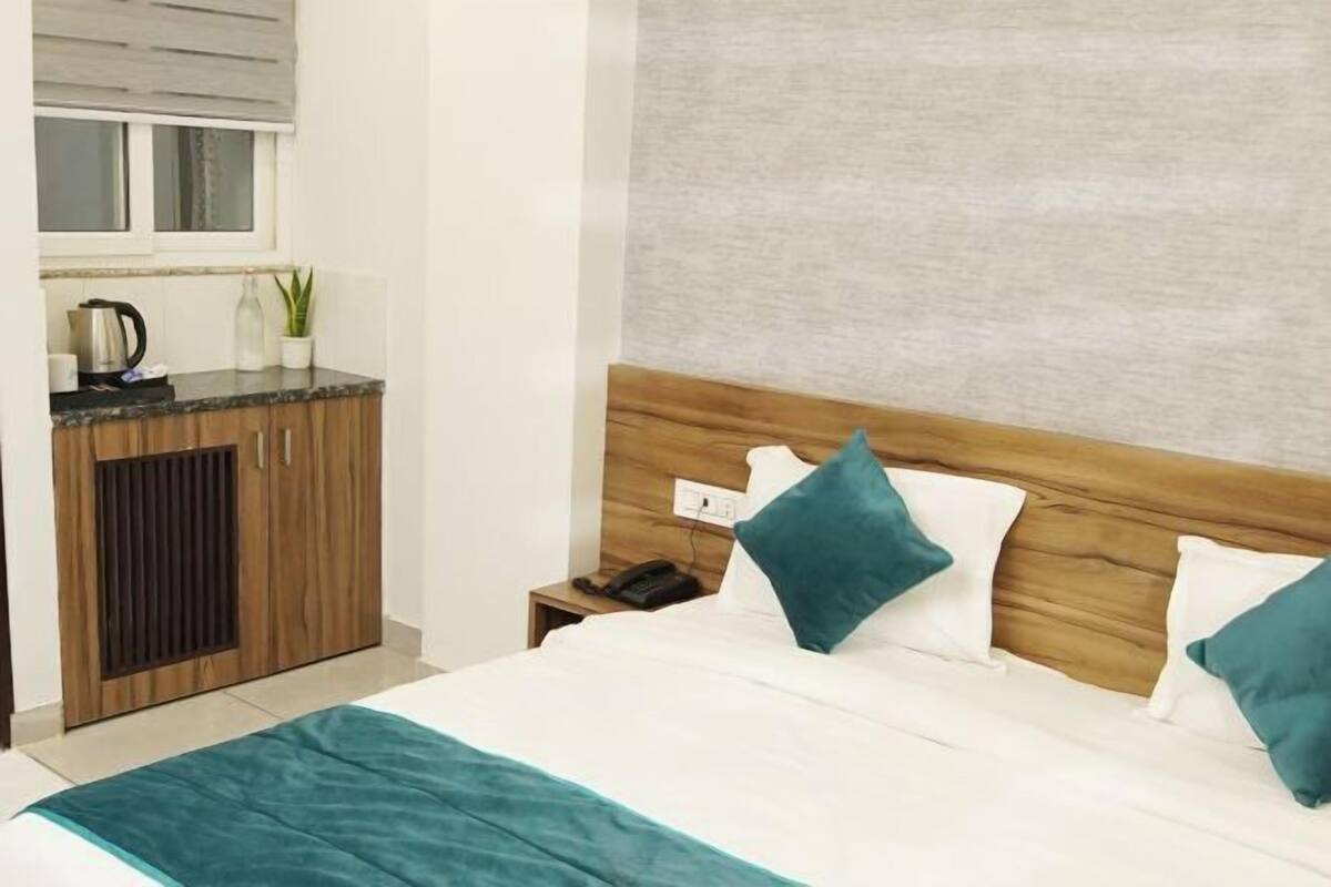 A hotel room with a bed, a wooden headboard, a nightstand, a telephone, a window with blinds, and a kitchenette area.