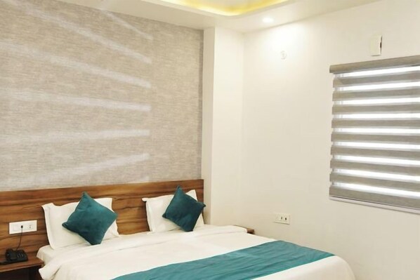 A hotel room with a bed, a nightstand, a wall-mounted light, and a window with blinds. - Gallivanto Inn (North Delhi)