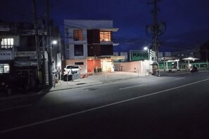 Vista de la calle al anochecer con un edificio moderno, una gasolinera y un coche aparcado.