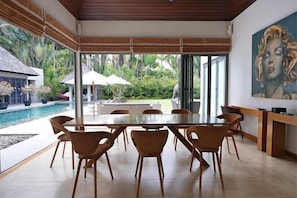 Un espace de repas moderne avec une table et des chaises en verre, une piscine et une vue sur un jardin tropical.