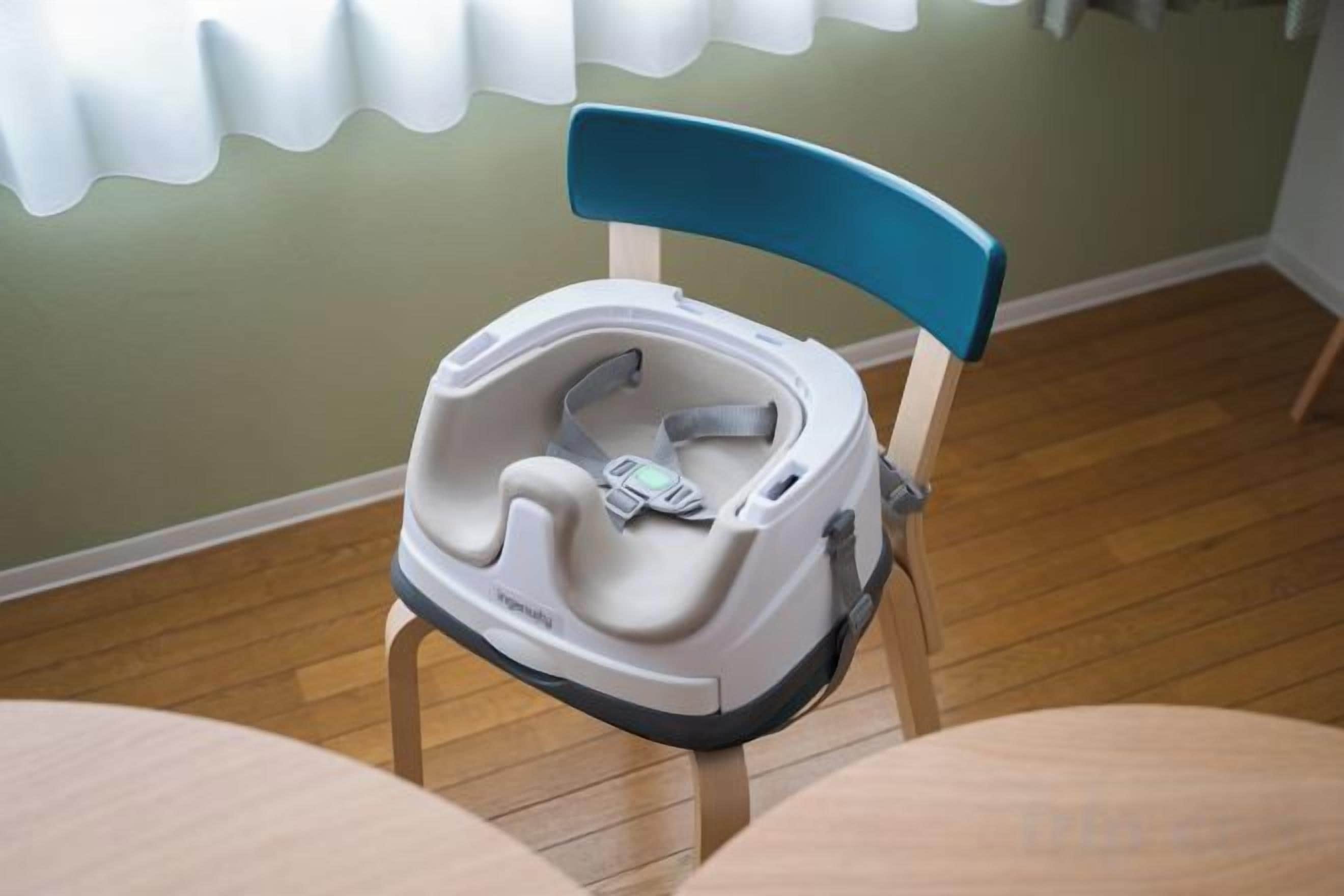 A high chair with a blue backrest and a tray, placed on a wooden chair in a room with a wooden floor and a green wall.