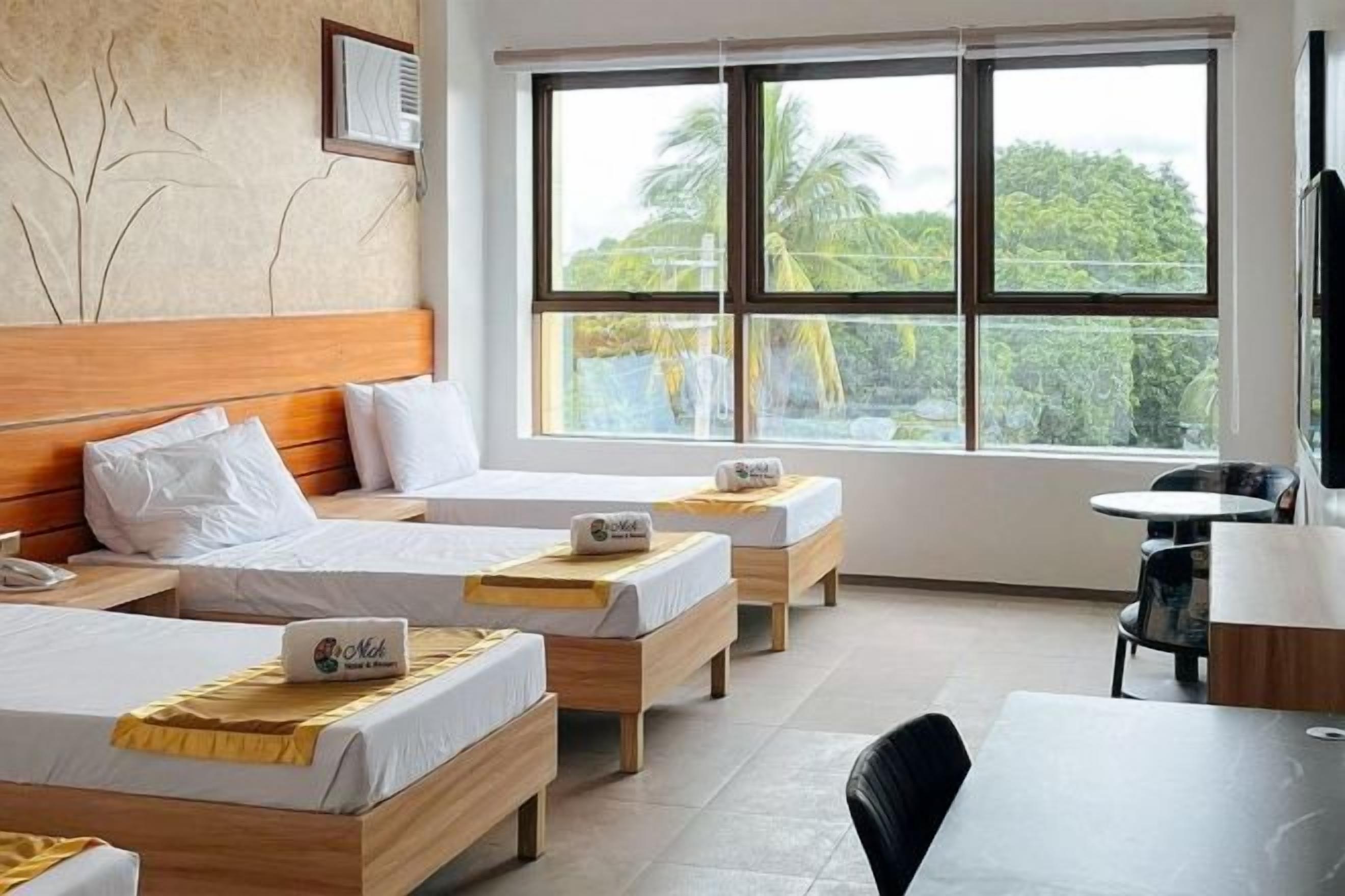 A hotel room with two beds, a wooden headboard, a small table, a chair, and a window with a view of greenery.