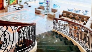 A hotel lobby with a modern design, featuring a staircase with ornate railings, a seating area with sofas and armchairs, and a large window offering a view of the street. - RedDoorz Plus @ Nick Hotel Guimba (Guimba)