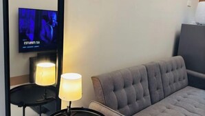 A room with a grey tufted sofa, two bedside lamps, a flat-screen TV mounted on the wall, and a small table with a lamp. - Solana Staycation Suites at Shore 3 Residence (Pasay)