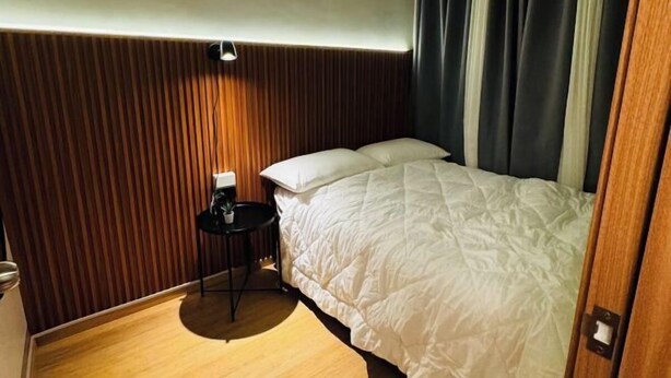 A hotel room with a wooden panel wall, a bed with white bedding, a small round table with a lamp, and a window with dark curtains. - Solana Staycation Suites at Shore 3 Residence (Pasay)