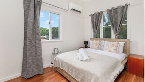 Une chambre à coucher comprenant un lit, des rideaux, un appareil de climatisation, une table de chevet et une fenêtre donnant sur un paysage verdoyant.