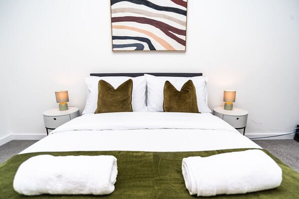 A neatly made bed with two pillows, a green throw, and two white towels. There are two bedside tables with lamps, and a piece of abstract art on the wall.