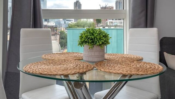 Uma área de refeições moderna com mesa de vidro rodeada por cadeiras brancas, um vaso de planta e jogos americanos trançados.