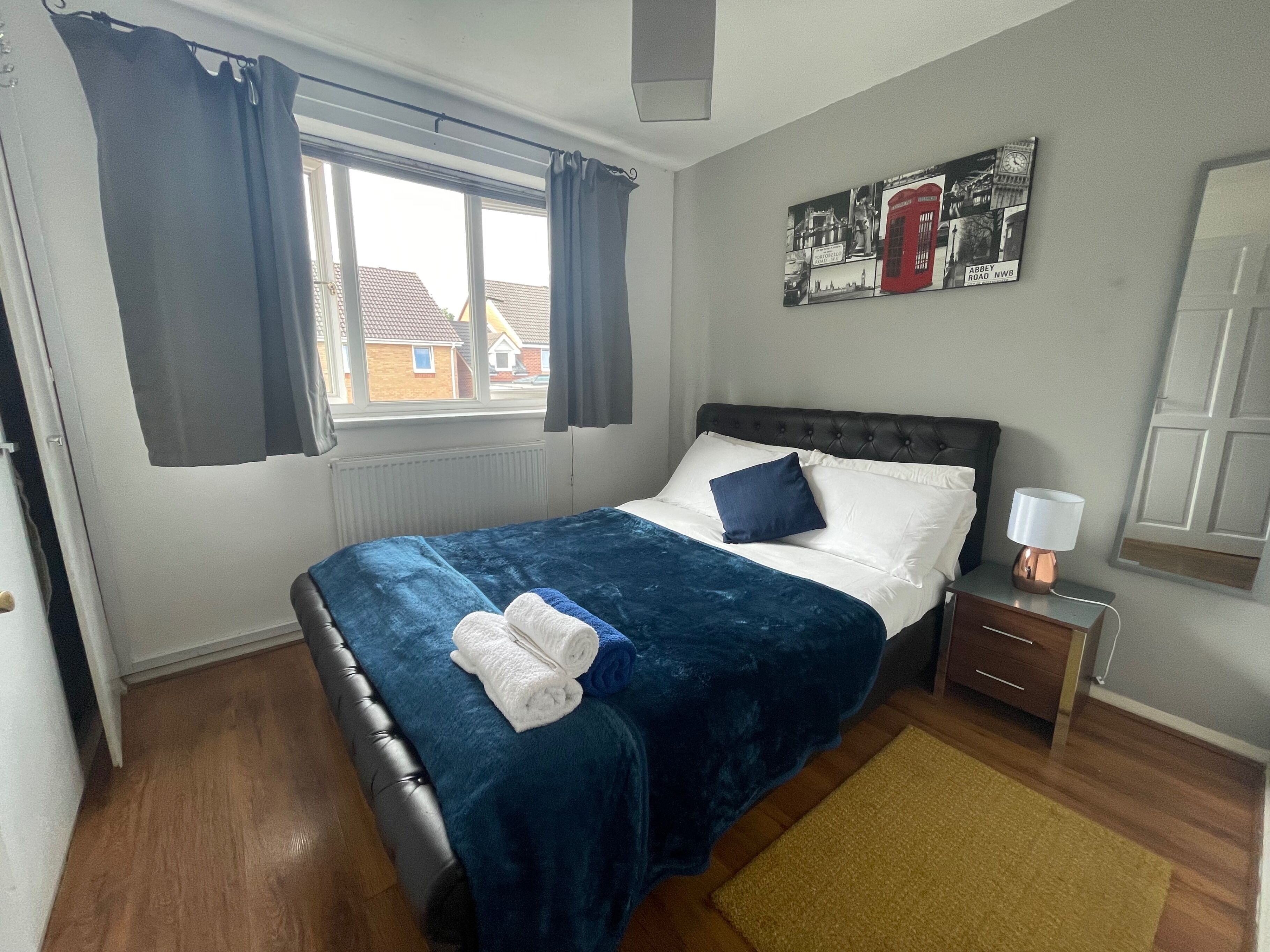 Een opgemaakt bed met een blauwe deken en kussens, een bedlampje en een lijst met een afbeelding aan de muur.
