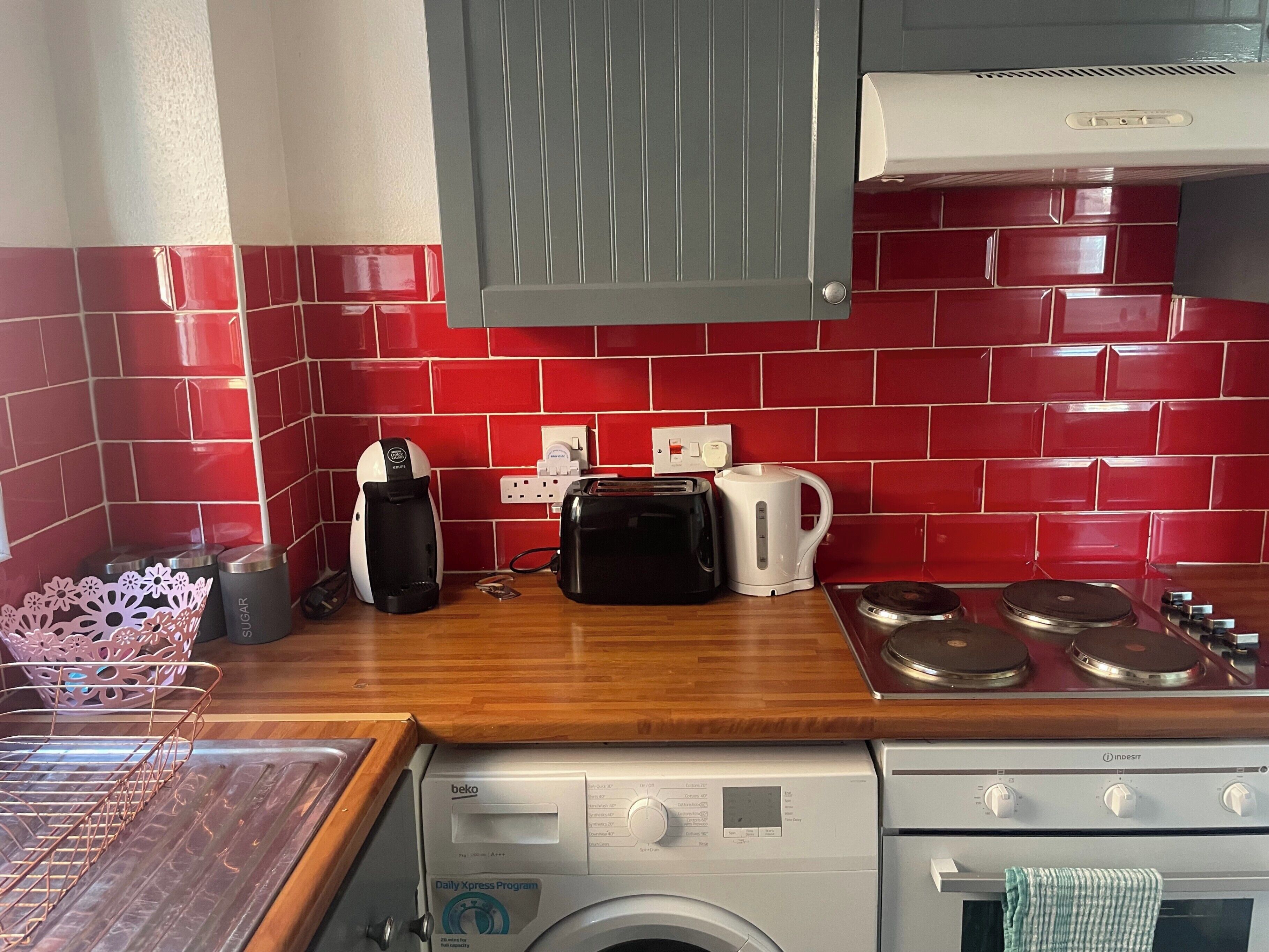 A compact kitchen with red tiled backsplash, a washing machine, a toaster, and a kettle.