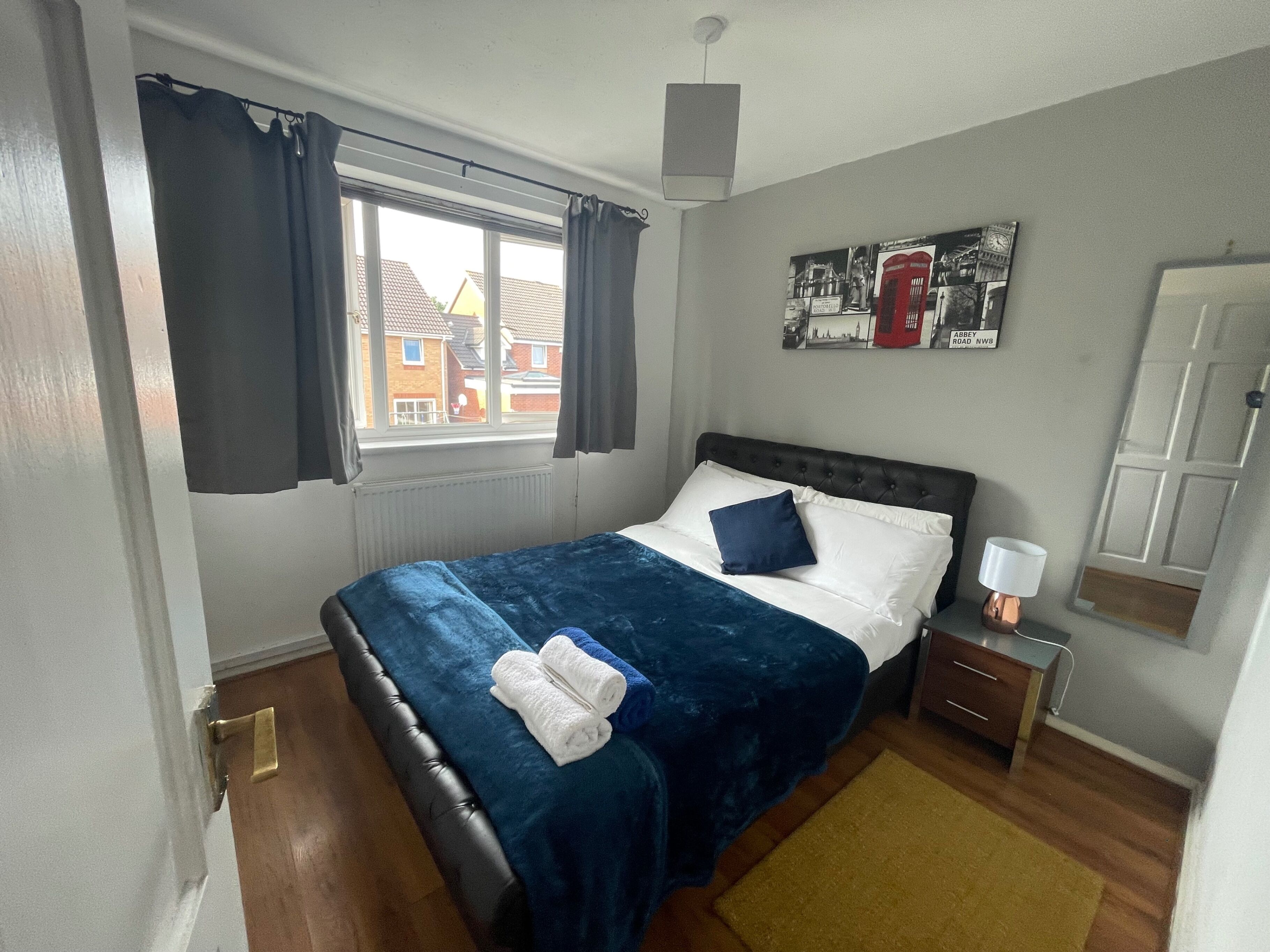 A bedroom with a bed, a nightstand, a lamp, a window with curtains, a radiator, and a wall with a picture.